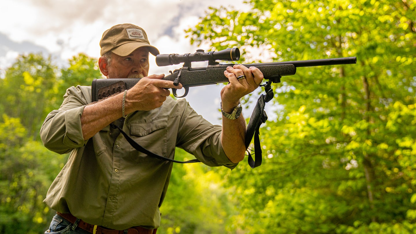 A shooter brings a rifle to his shoulder on a backyard range with green foliage in background.