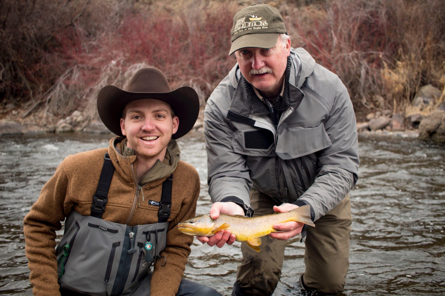 A young fisherman in a cowboy hat stand stands to an older fisherman holding a brown trout