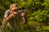 A hunter shoots a rifle with a suppressor off from shooting sticks, with green foliage in background.