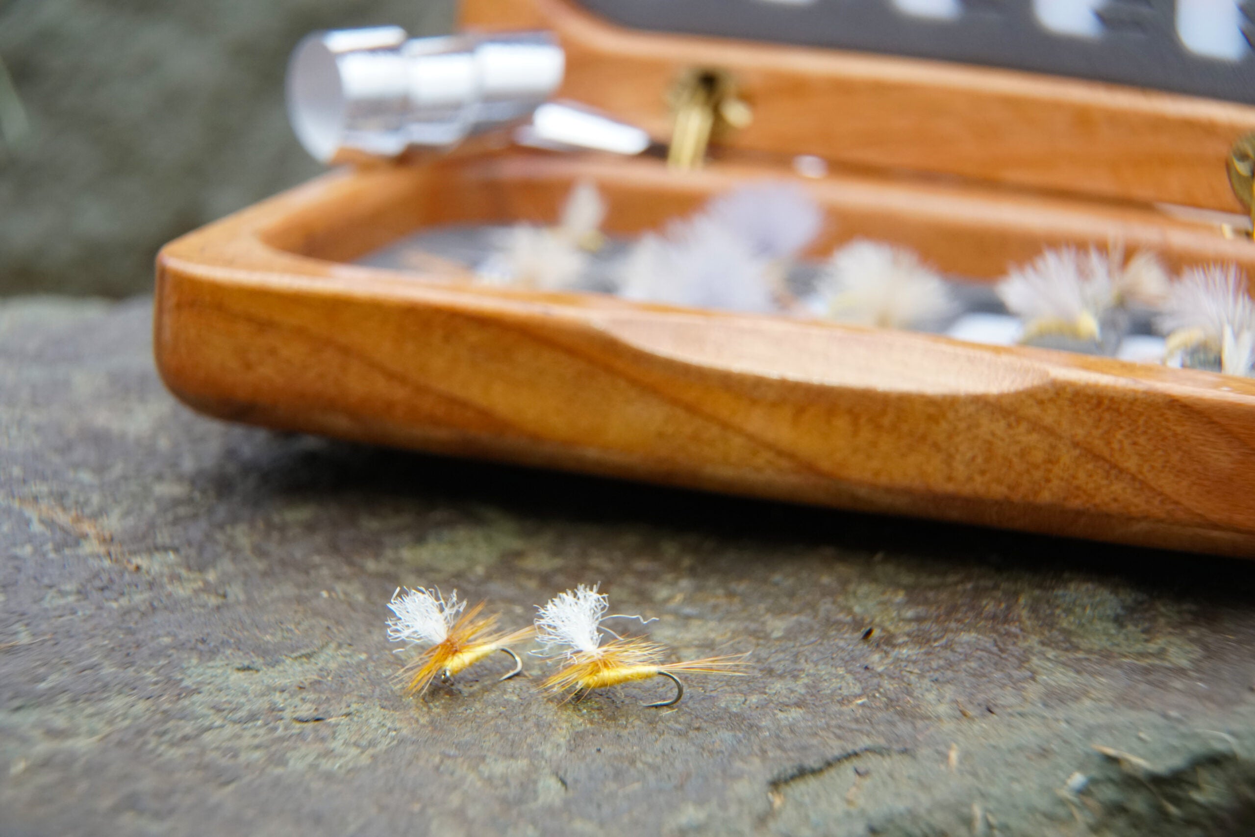 Two dry flies on a rock with a fly box in the background