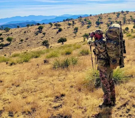 Hunter in camouflage gear using binoculars on arid grassland hillside with mountains.