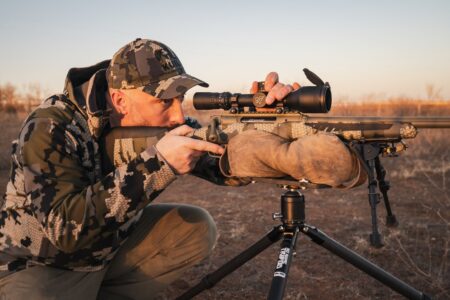 Hunter in camouflage aiming precision rifle with scope on bipod at sunset.