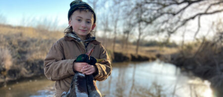 Young child in camo hat holds harvested mallard duck near rural creek