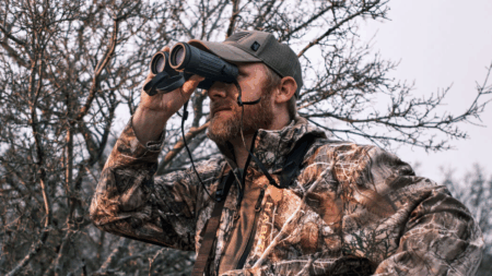 Hunting Gear Is Up to 50% Off at Cabela’s Right Now Man in camouflage jacket using binoculars outdoors during hunting season