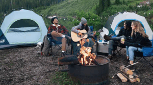 Four friends relaxing around a campfire with tents and guitar in nature.