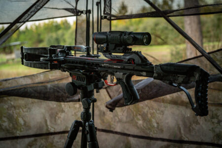 Black tactical crossbow mounted on tripod with scope in outdoor hunting blind