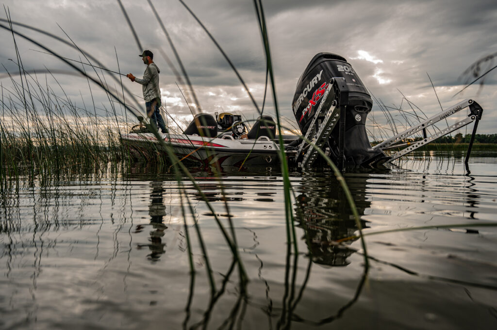 Angler fishing from Mercury-powered bass boat near reeds on calm lake at dusk