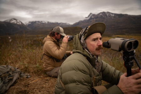 Two hunters using binoculars to scout wildlife in a mountainous wilderness landscape.