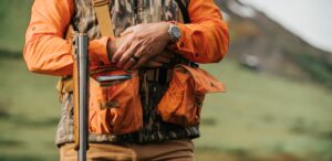 Hunter in orange safety vest holding rifle in mountain landscape