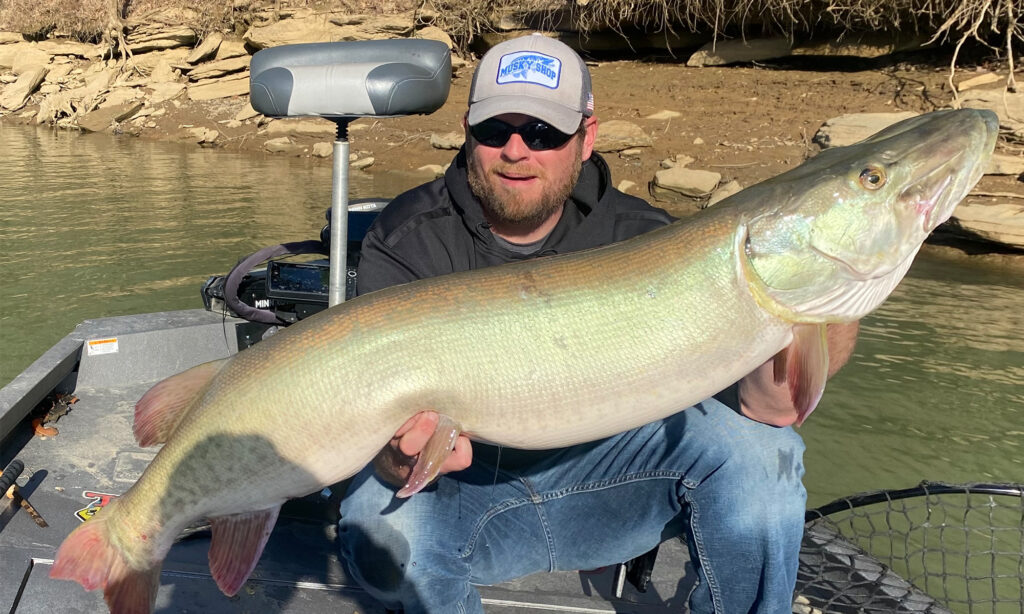 Angler holding large muskie fish on boat near rocky riverbank