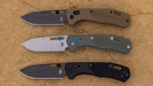 Three Gerber folding pocket knives in tan, green, and black on stone surface.