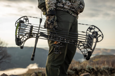 Bow Bootcamp: Get Your Compound Bow Ready for the Season Hunter in camouflage carrying a compound bow with arrows in outdoor terrain.