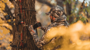 Hunter in camouflage checking phone beside compound bow leaning against tree.