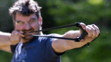 The Best Slingshots Man aiming a black slingshot outdoors with green trees in background