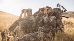 Two camouflaged bow hunters scanning open grasslands with binoculars during hunting season.