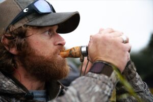Bearded hunter in camouflage blowing a wooden duck call while wearing sunglasses