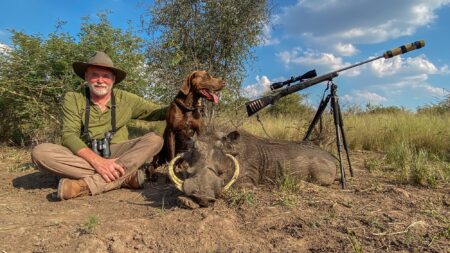 Hunter poses with harvested warthog, hunting dog, and scoped rifle outdoors.