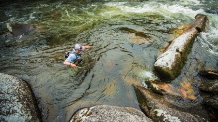 Fly fisherman wading through rocky river rapids holding fishing rod