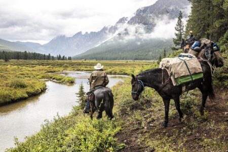 Two riders on packed horses pause beside a mountain stream in misty wilderness.
