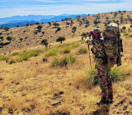 Hunter in camouflage using binoculars on dry mountain hillside with scenic valley view.
