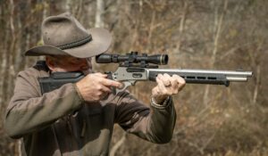 Man in cowboy hat aiming scoped lever-action rifle outdoors in wooded area