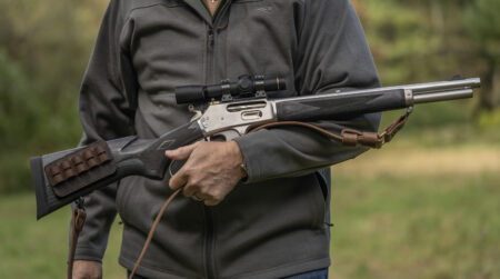 Carry Strap vs Sling: Which Should You Put on Your Rifle? Person holding a lever-action rifle with scope and leather sling outdoors.