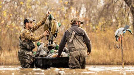 Two hunters in camo waders collecting duck decoys from a boat in shallow water.