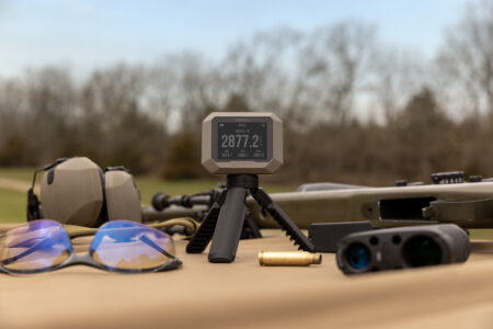Garmin shooting chronograph displaying 2877.2 fps beside rifle, ear protection, and safety glasses