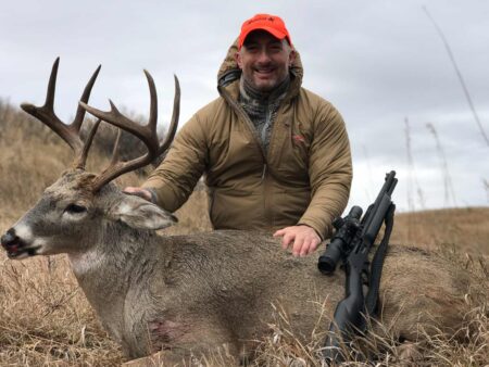 A Love Letter to the Lever-Action Hunter in blaze orange cap poses with harvested whitetail buck and rifle outdoors.