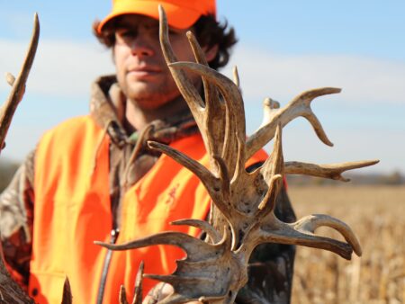 Hunter in blaze orange holds large non-typical whitetail deer antlers in field.