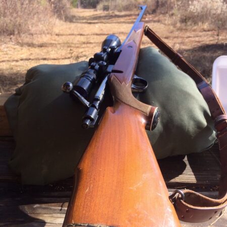Scoped hunting rifle with wooden stock resting on green shooting bag outdoors