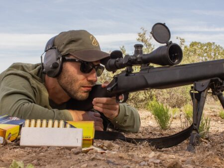 Best Long-Range Hunting Cartridges of 2025 Man in cap and ear protection aims scoped rifle prone at outdoor shooting range.
