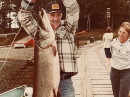 Man in CAT cap proudly holds large caught fish on wooden dock.