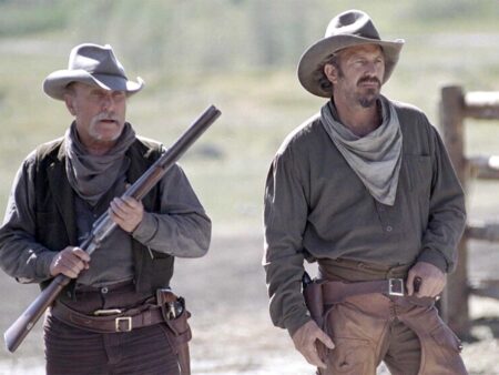 Two armed cowboys in western attire walk through a dusty frontier landscape.