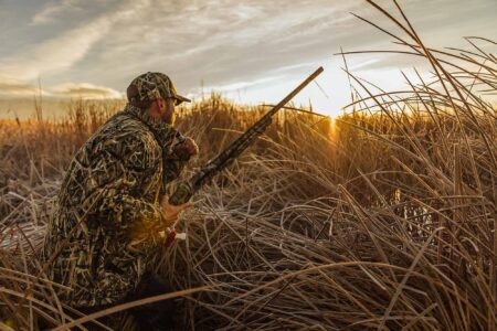 Hunter in camouflage gear holding shotgun in tall marsh grass at sunset.