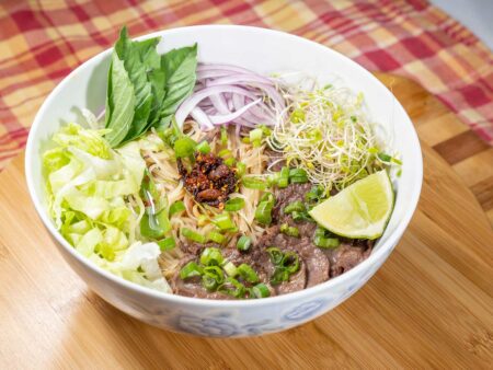 Bowl of Vietnamese beef pho with fresh herbs, rice noodles, and lime wedge.