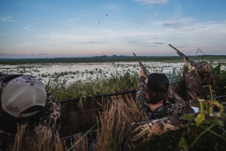 Hunters in camouflage aiming shotguns at ducks over a marsh at dawn