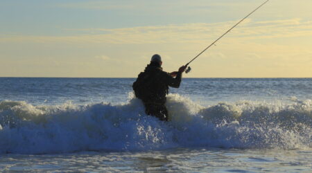 Fisherman surf casting in ocean waves at sunset wearing waders and backpack
