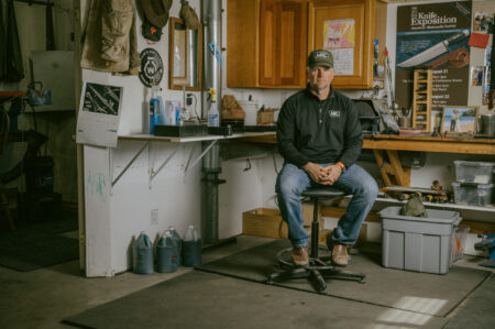 Man in MKC cap sits in workshop with knife exposition poster behind him.