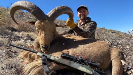 Hunter posing with harvested aoudad ram and rifle in desert terrain.