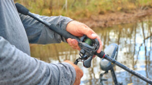 Angler holding baitcasting fishing reel and rod near calm water outdoors