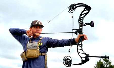 Man aiming compound bow outdoors wearing Leupold vest and baseball cap.