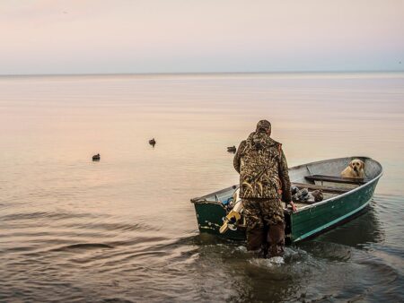 Duck hunter in camouflage pushing boat into calm lake with Labrador retriever aboard.