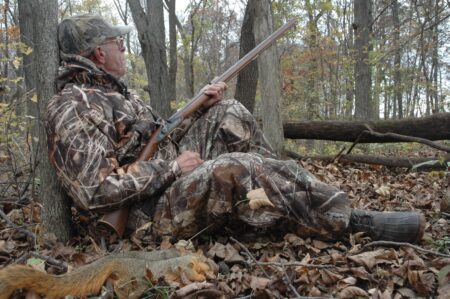 Hunter in camouflage gear resting against tree in autumn forest holding rifle.