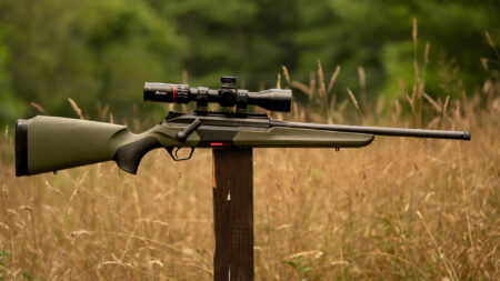 Green bolt-action rifle with scope resting on wooden post in field