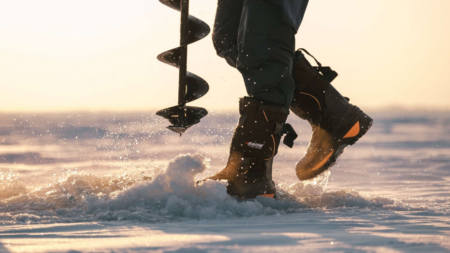 Person using ice auger drill while wearing Baffin winter boots on frozen lake.