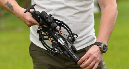 Person holding a black compound crossbow with orange accents outdoors.