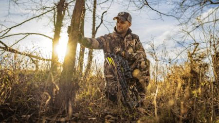 Cabela’s Is Having a Massive Sale on Archery and Bow Hunting Gear Right Now Hunter in camouflage crouching in woods holding compound bow at sunset.
