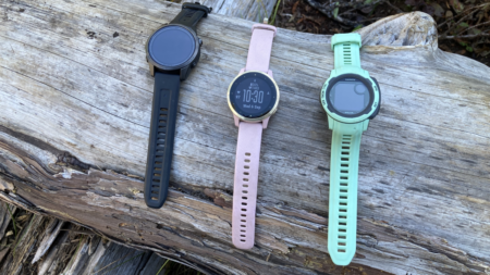 Three Garmin smartwatches in black, pink, and mint green on wooden log.
