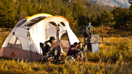 Family camping in mountain meadow with large tent, chairs, and portable grill.